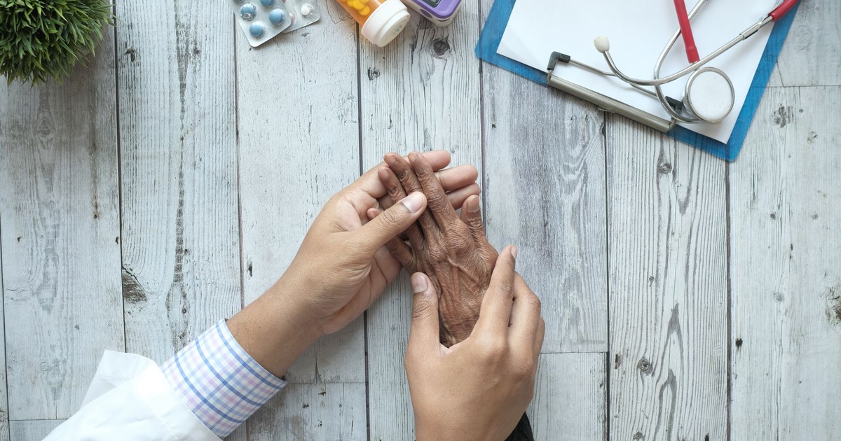 A doctor holding an elderly patient's hands on a desk with medication and stethoscope.
