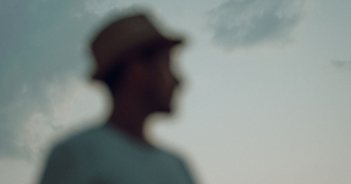 A blurred silhouette of a man wearing a hat, captured during twilight with a serene sky as background.