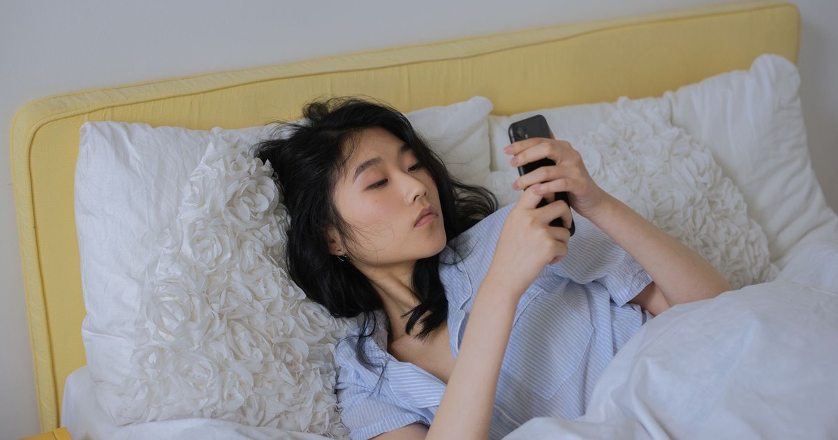 Asian woman comfortably lying in bed, using her phone in a serene bedroom setting.