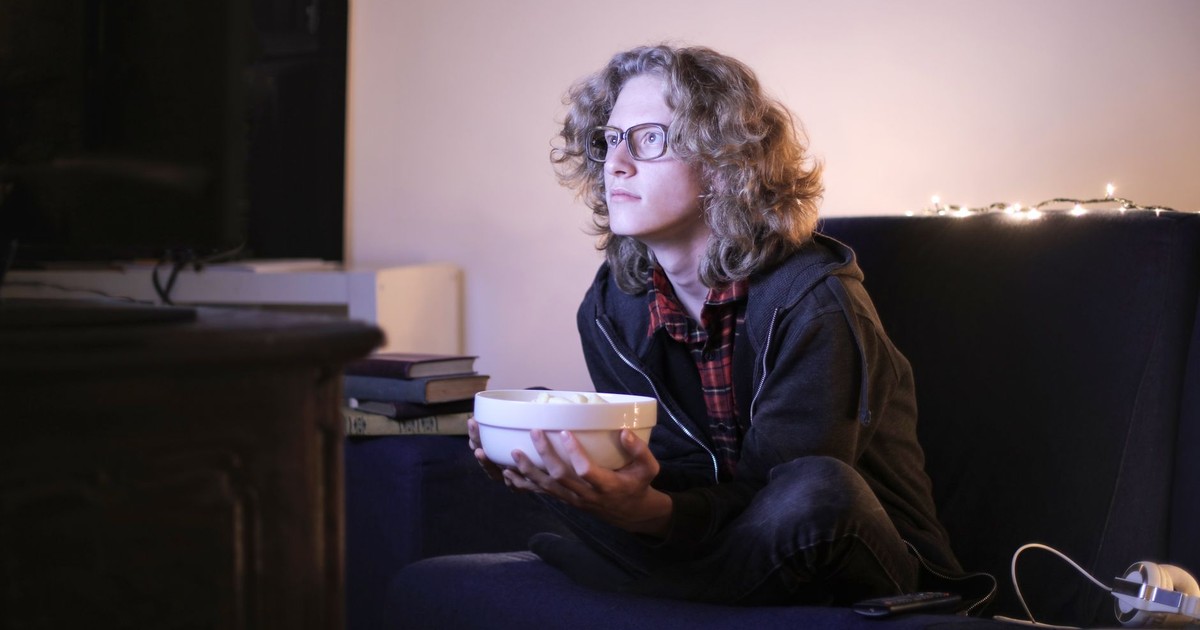 Serious young male with long curly hair sitting with crossed legs on couch in living room and enjoying interesting movie while chilling at home during weekend