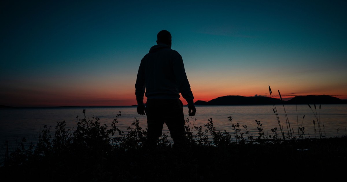 A solitary man stands by the water's edge, silhouetted against a vibrant sunset.