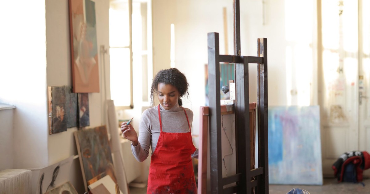 Young black lady in casual clothes and red dirty apron standing near easel with canvas holding paintbrush in hand and looking down while creating artwork in workshop