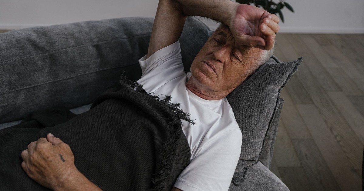 Elderly man relaxing on a sofa, hand on forehead, white shirt and blanket.