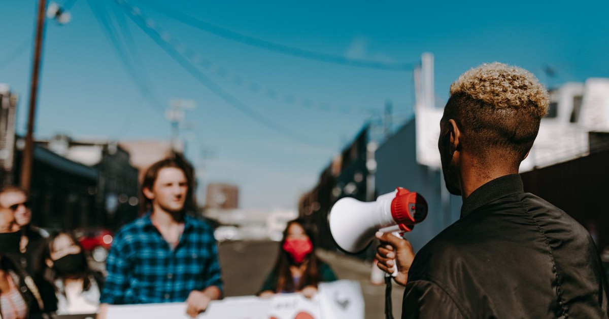 A diverse group of people protesting in an urban setting, with a focus on a speaker using a megaphone.