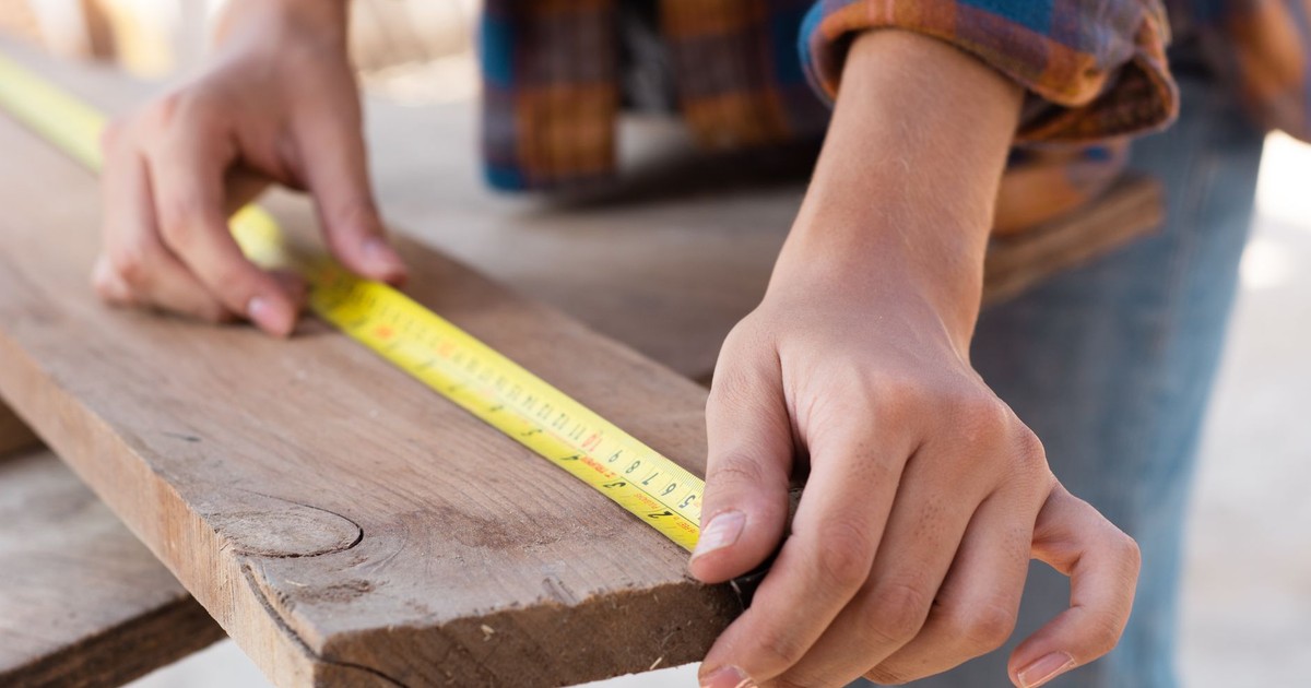 Close-up of hands measuring a wooden plank with a tape measure in a carpentry setting.