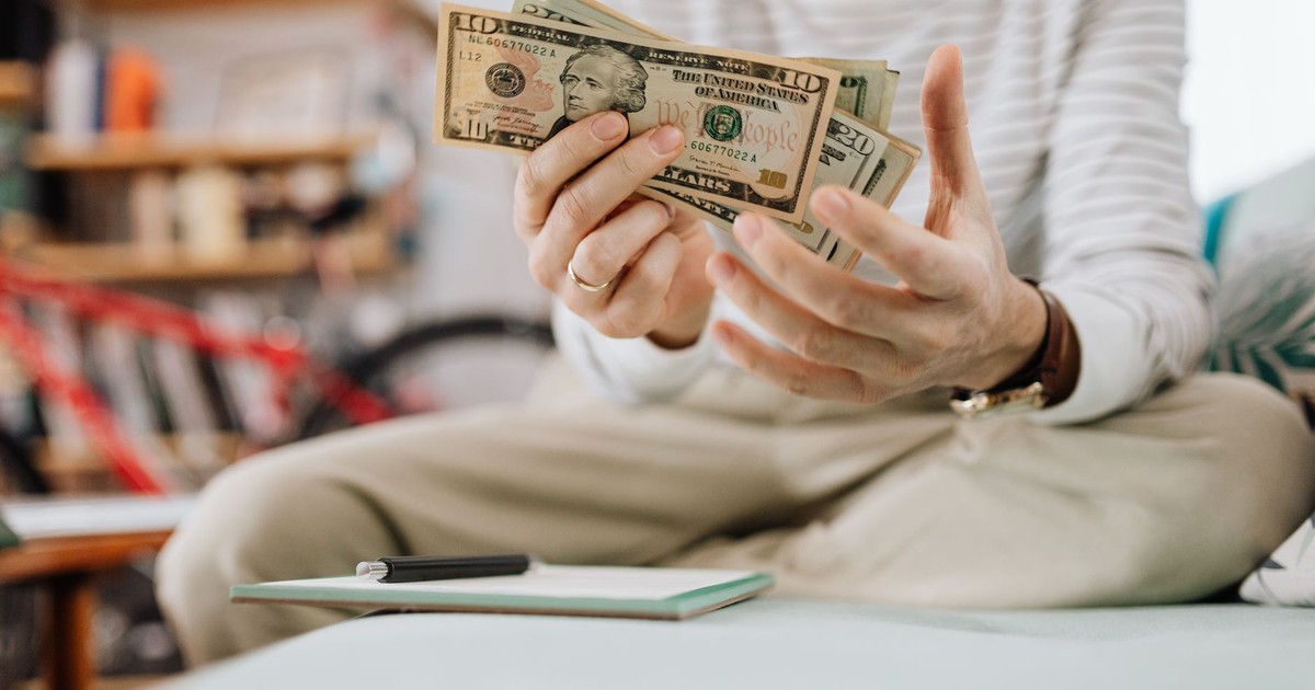 Man wearing casual clothes counts dollar bills while sitting on a sofa indoors.
