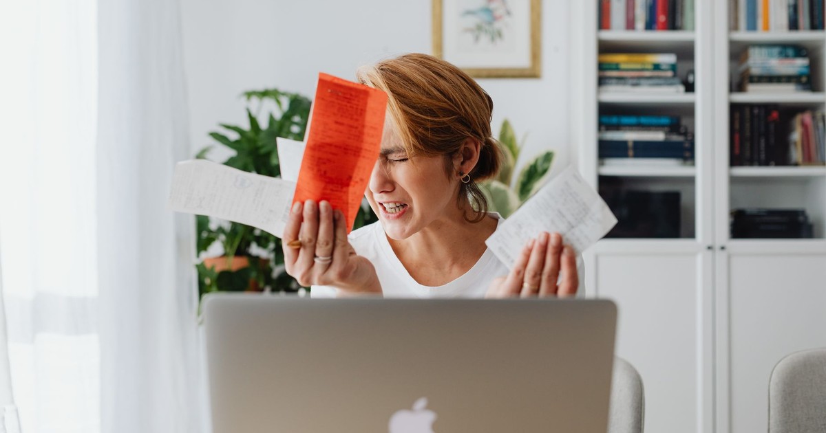 Frustrated woman managing finances, surrounded by bills and a laptop, looking distressed.