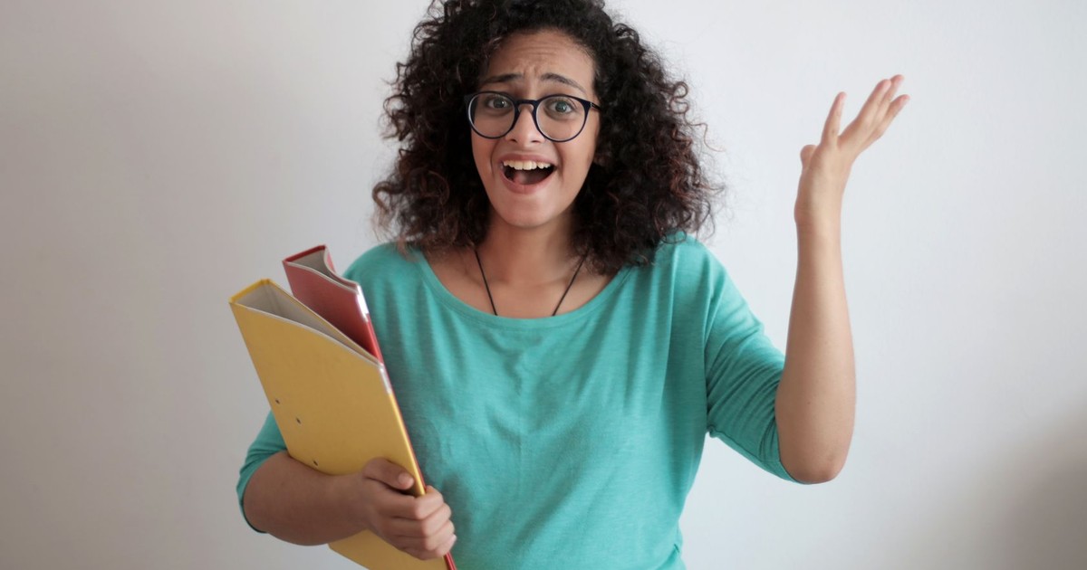 Dissatisfied annoyed woman with mouth opened wearing glasses and turquoise blouse looking away and screaming while standing against white wall with folders of documents and having problems in work