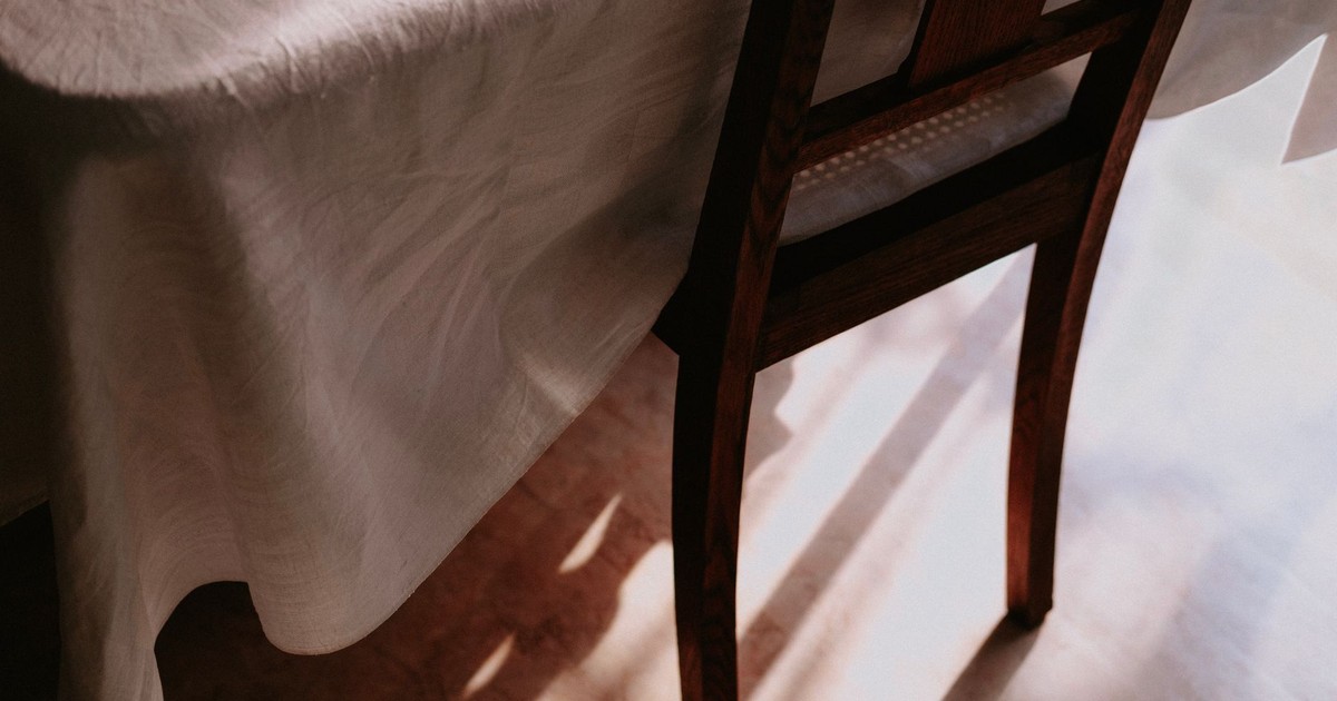 A wooden chair stands beside a table draped with a white tablecloth, bathed in warm sunlight indoors.