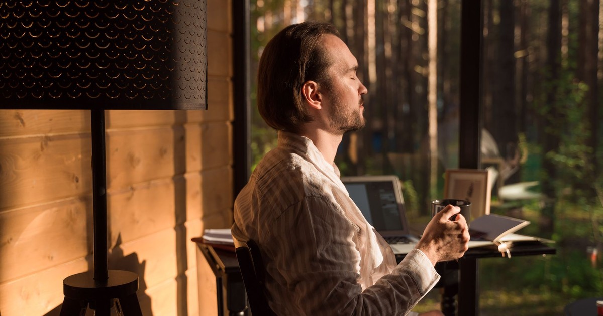 A man enjoying coffee while working remotely in a sunlit cabin, surrounded by nature.