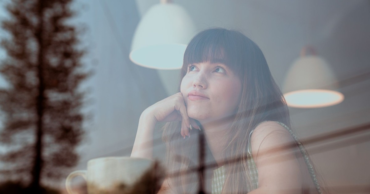Thoughtful woman gazing outside a café window with reflections and warm lighting.